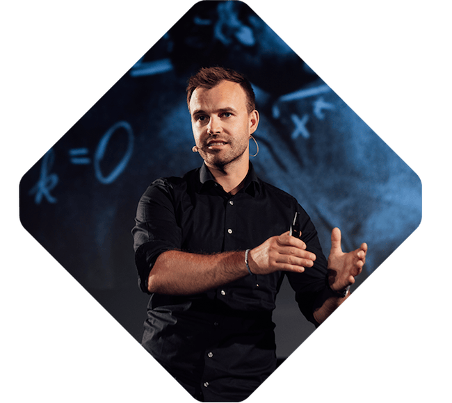 A man in a black shirt speaks into a headset microphone while gesturing with his hands, with mathematical equations visible on a screen behind him during the Spryker EXCITE 2023 event.