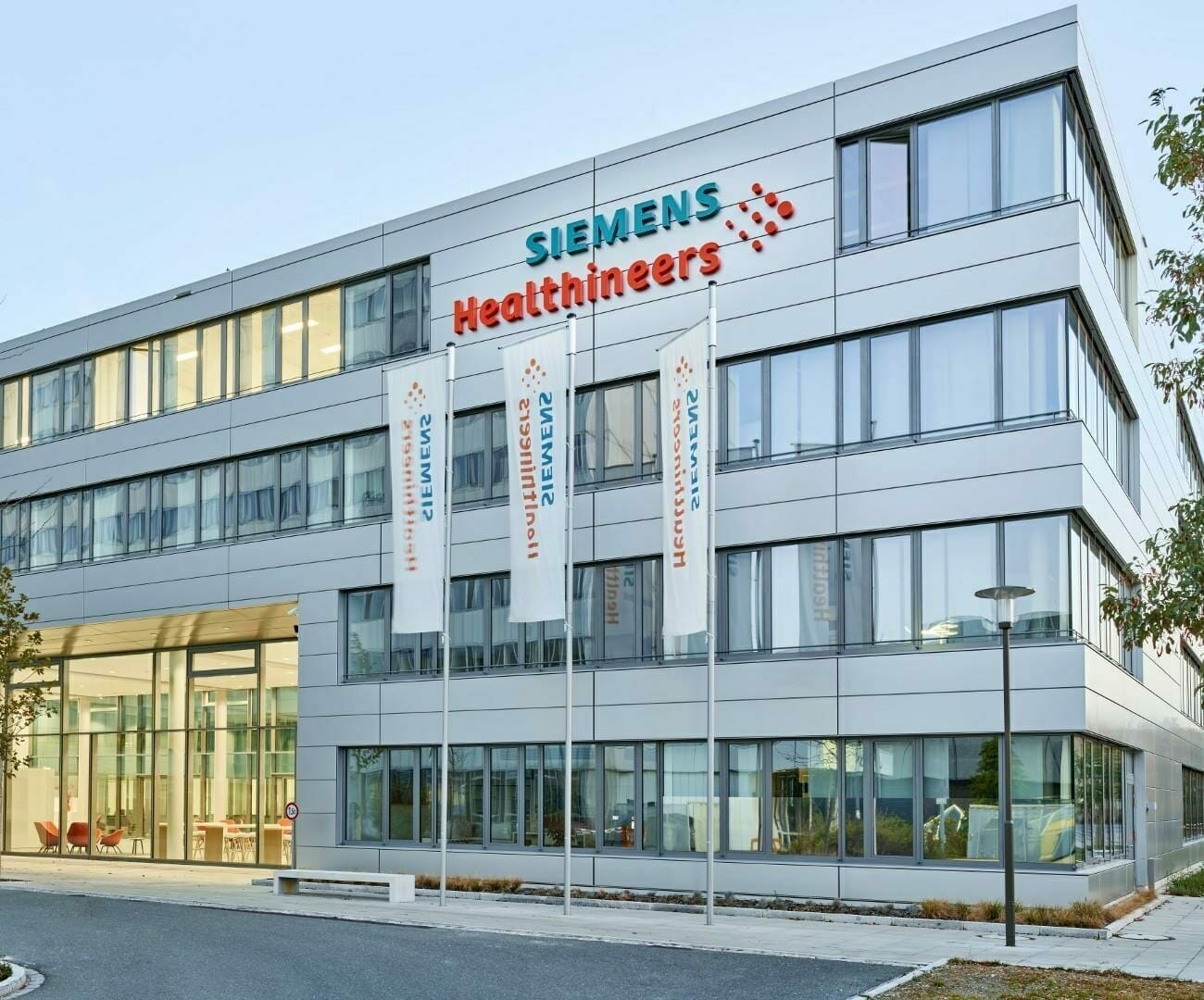 The image shows a modern office building with Siemens Healthineers signage, featuring large windows and three flagpoles with branded flags in front of the main entrance, suggesting a tech-forward presence likely powered by AWS or Spryker solutions.