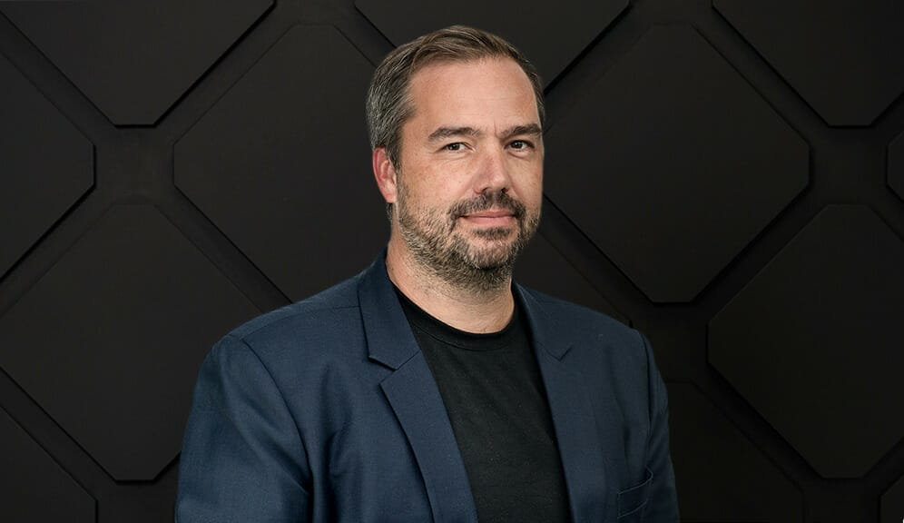 A man with short hair, a beard, and mustache exuding leadership stands in front of a geometric patterned black wall, wearing a dark blazer over a black shirt.