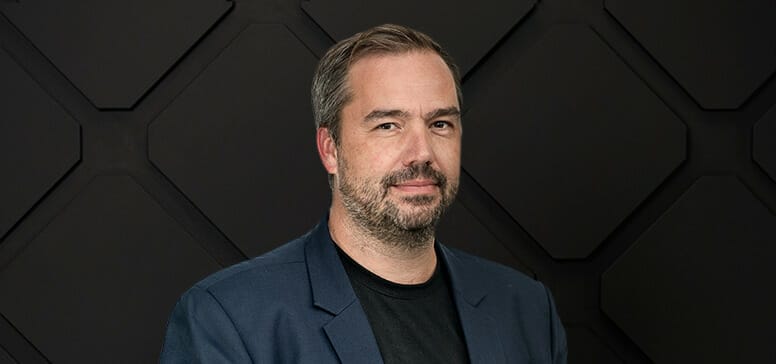 A man with short hair and a beard, wearing a dark blazer and black shirt, stands against a geometric black background, exuding an air of leadership.