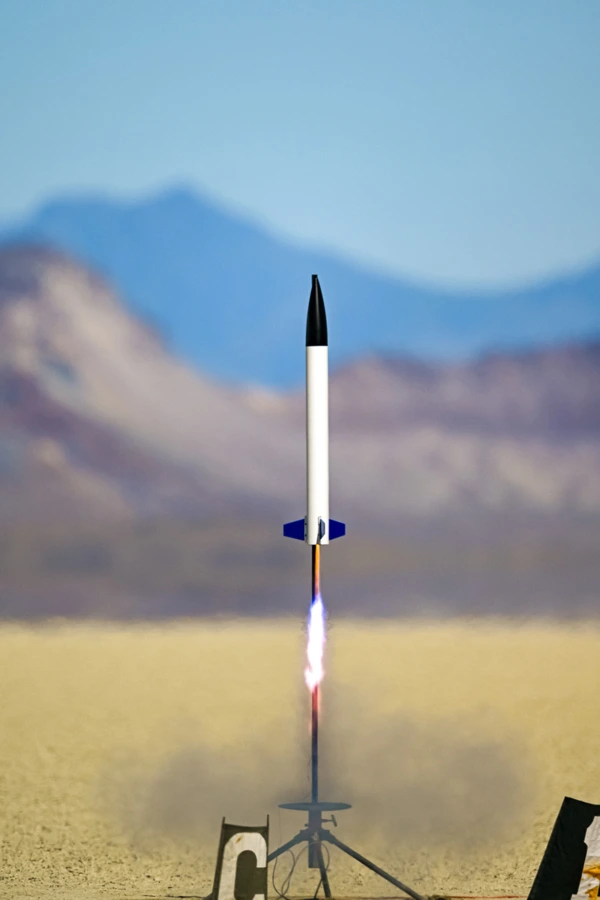 A rocket with a white airframe, black nose cone, and blue fins launches off a rail with desert and mountains in the background. The motor propelling it has a long blue flame