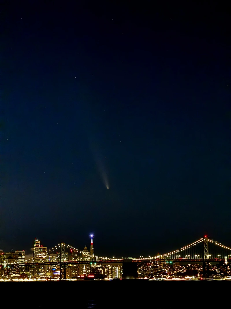 A comet photographed from looking west towards the Bay Bridge. The comet is mid-frame, with a long tail pointing upwards, seen between two of the towers of the San Francisco side of the bridge. There is a bright blue light atop the Transamerica Pyramid building to the left.