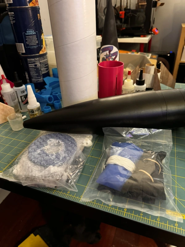 The unboxed kit on a work table with the airframe tube, the nose cone, a package with the machined motor mount plates, fins, and hardware, and another package with the recovery gear.