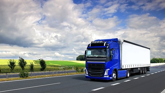 Large eighteen-wheeler driving on a road in an overcast day with cornfields in the distance.