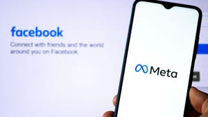 A phone with the meta logo on the screen held up in front of a background with the original Facebook login page