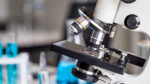 Microscope examining slide of sample with test tubes in background.