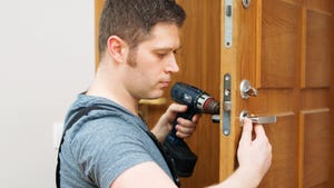 Young handyman in uniform, overalls and a gray t-shirt, changing a door lock using an electric drill.