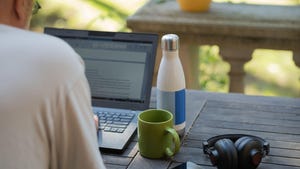 Man writing on laptop on a patio, with coffee mug and headphones in reach