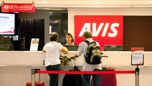 Two people standing at an Avis car rental counter speaking to an employee behind the desk