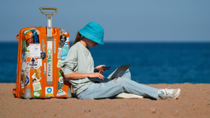 Woman in blue hat, blue jeans and a sweatshirt using a laptop and mobile device sitting on a beach against an orange roller bag with stickers on it