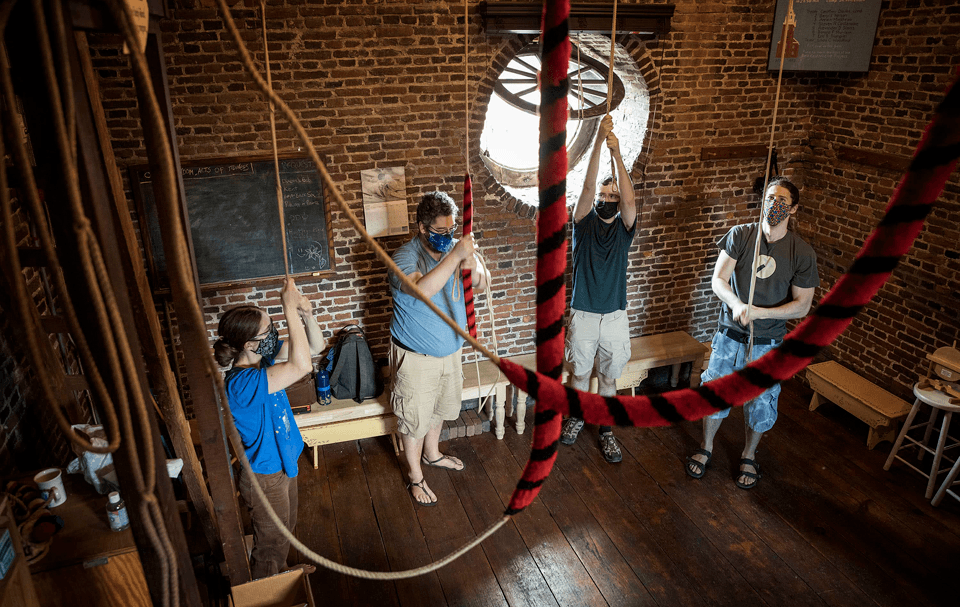 Photo of one woman and three men wearing masks pulling on ropes inside a brick structure with a ground window opened in the background