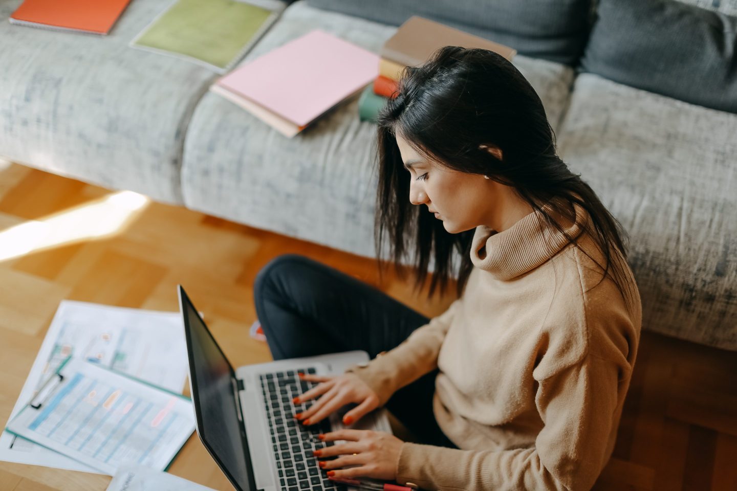 Woman sits criss-cross next a couch while working on a laptop.