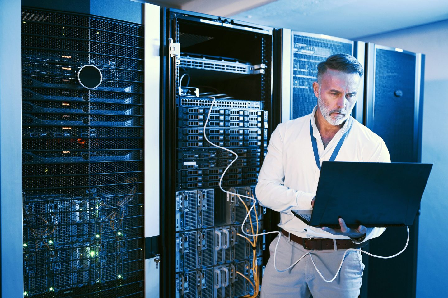 Man works in a computer network room, holding his laptop while standing up