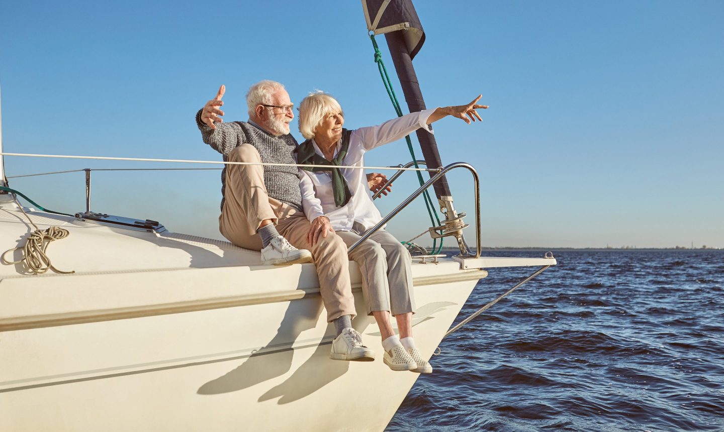 A happy senior couple sitting on the side of a sail boat on a calm blue sea, pointing at landscape, enjoying view. Sailing, age, tourism, travel and people concept. Horizontal shot