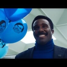 Milchick from "Severance" smiling while holding blue balloons with Mark S.'s face on them.