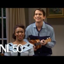 Black woman in blue dress sitting down. Behind her is a white man in grey button up playing a ukulele