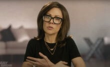 A woman in a brown wig and glasses speaks into the camera.