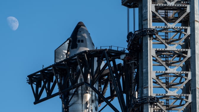 A large metal structure holds up the Space X Starship against a blue sky.