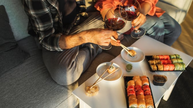 Two people sitting on couch holding wine glasses and eating sushi