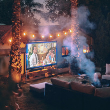 Screen on side of house surrounded by fairy lights showing video and facing set of couches around a fire