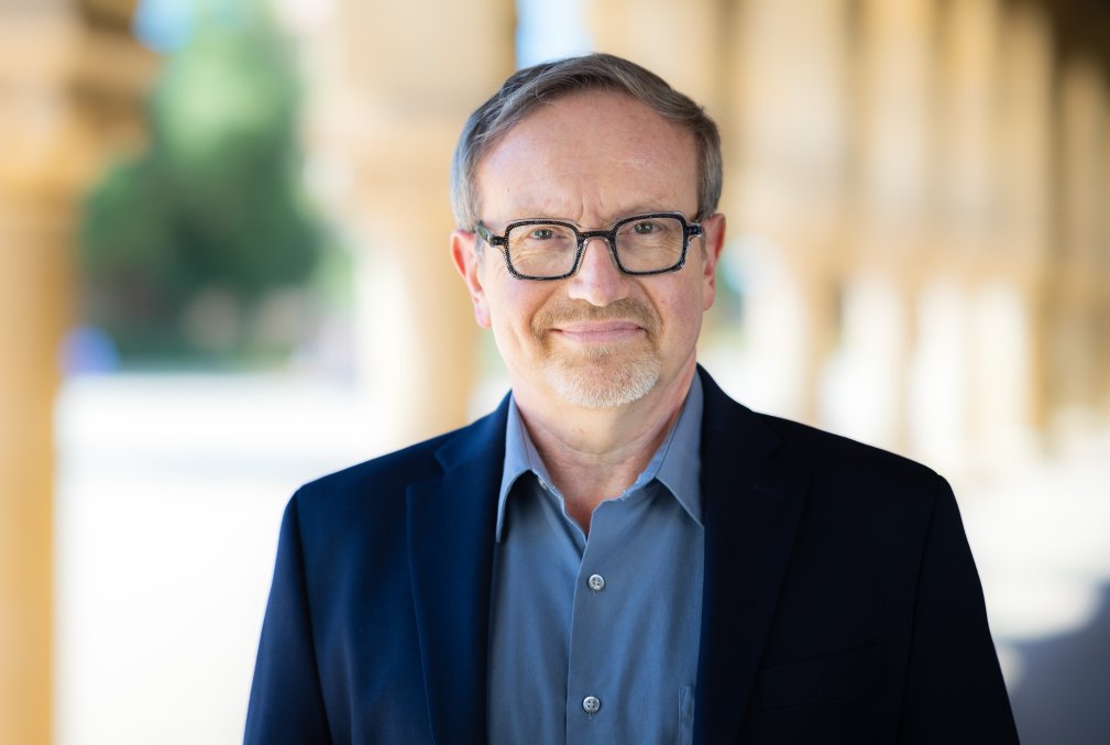 Aron Rodrigue wearing glasses, a blue collared shirt, and a navy-blue blazer standing in one of the arcades on Stanford's Main Quad