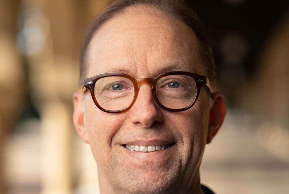 Russell Poldrack wearing glasses, a light-blue collared shirt, and a navy blazer standing in one of the arcades along Stanford's Main Quad