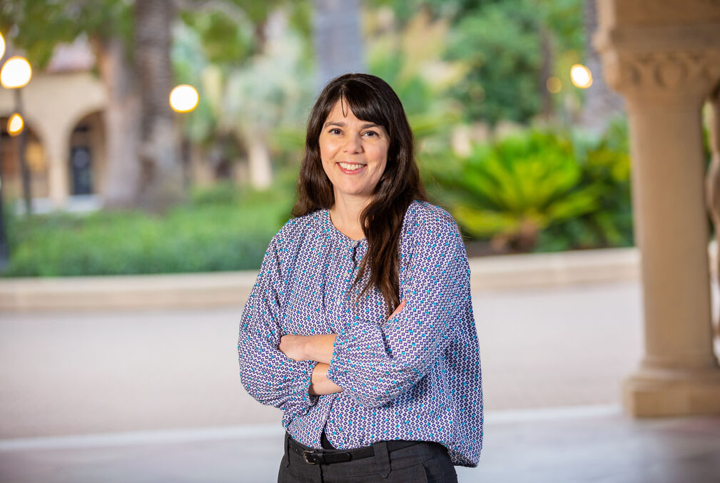 Kristy Red-Horse wearing a light-blue blouse and standing with arms crossed under one of the arcades along Stanford's Main Quad