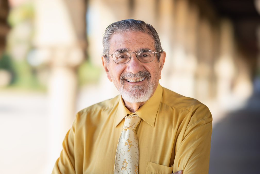 Richard Zare wearing a mustard yellow shirt and tie standing under one of the arcades on Stanford's Main Quad