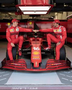 two men in red racing suits sitting on the back of a race car with their arms around each other