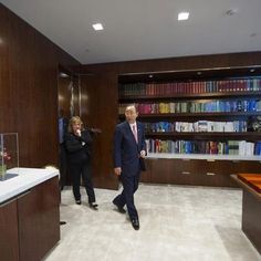 two people are walking in an office with bookshelves and desks behind them