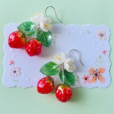 three cherries with leaves and flowers are hanging from earrings on a doily card