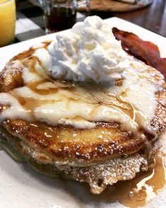 a stack of pancakes topped with whipped cream and syrup on a plate next to bacon