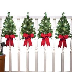 four christmas trees with red bows tied to them on a white railing next to a potted plant