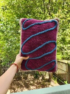 someone holding up a crocheted pillow in front of a fenced area with trees