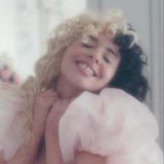 a woman with curly hair wearing a white dress and smiling at the camera while holding her hands on her chest