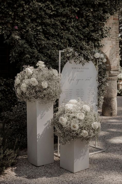 White roses and white gypsophila with white wedding table plan signage White Rose Wedding Arrangements, White Flower Table Arrangements Wedding, White Flowers Arrangements Wedding, White Flower Wedding Backdrop, White Table Flower Arrangements, White Flowers Decoration Wedding, Wedding Decorations White Roses, All White Floral Arrangements Wedding, White Rose Isle Wedding