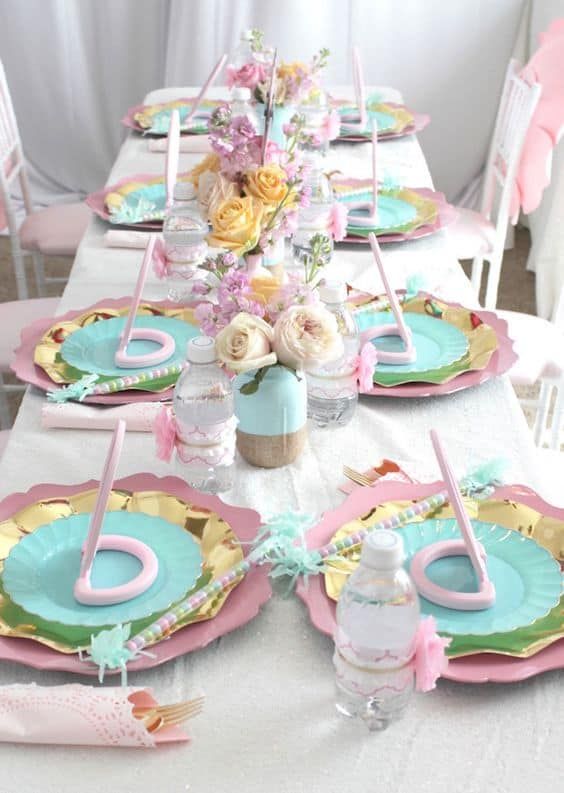 the table is set with pink and blue plates, silverware, flowers and napkins