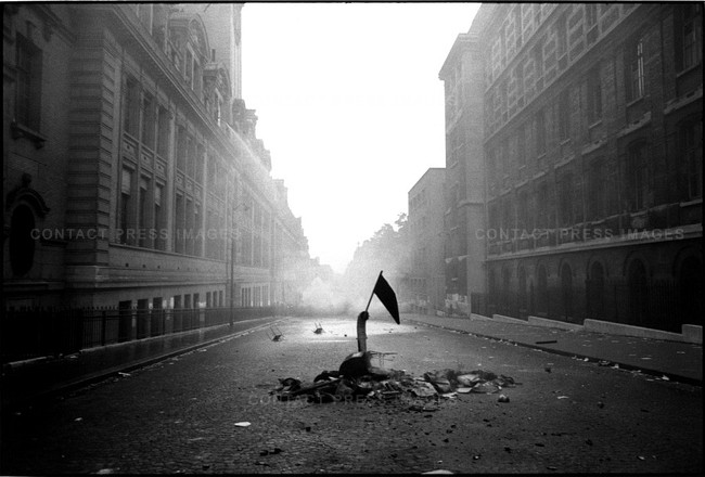 A Black & White picture oh a hand graspind a small black flag, rising from an erupting steet.