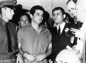 A Black & White Picture of a Man in handcuffs, surrounded by police and politicians of the Greek Junta