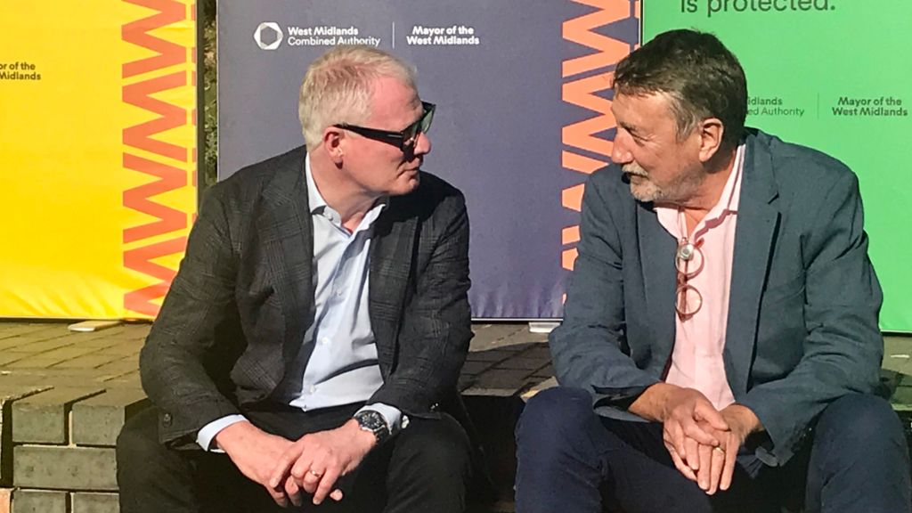 West Midlands Mayor Richard Parker and Steven Knight sat on a brick wall in front of hoardings, both wearing suits and speaking to each other