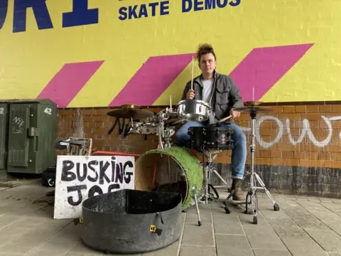 Joey Swindells, a 28-year-old busker from Bolton, sits at his makeshift drum set. His hand-written "Busking Joe" sign is propped up in front of him, next to his equipment case 