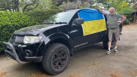 A man in khaki t-shirt and shorts leans on the side of the black Mitsubishi Warrior car, a big 4x4 with a grill on the front, high wheels and a Ukraine flag draped across one of the side windows. It is parked on a road with a bush behind it.