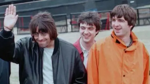 PA Media A photo of Oasis at Knebworth in 1996, Liam is wearing a leather jacket and waving. Noel is wearing an orange coat and smiling.