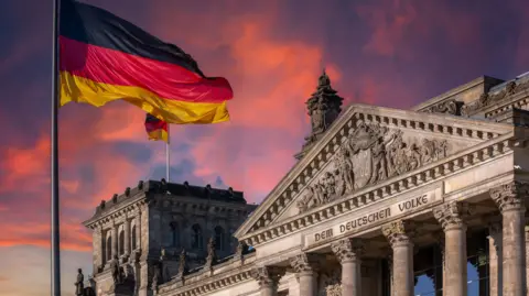 The German parliament building with a German flag fluttering in the foreground, and a dramatic pink sunset in the background