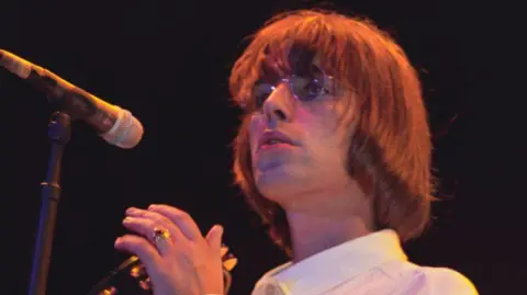 Getty Images Liam with long hair holding a tambourine, wearing round glasses reminiscent of John Lennon and standing in front of a microphone