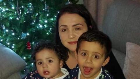 A close-up of a woman and two children. They are facing the camera, and there is a Christmas tree behind them. 