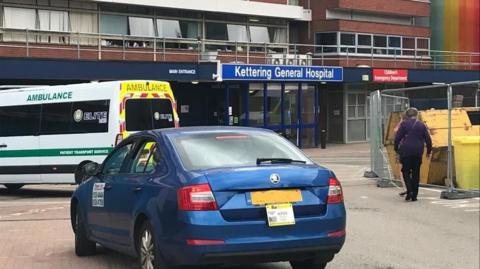 The entrance to Kettering General Hospital with an ambulance parked outside and blue car. 