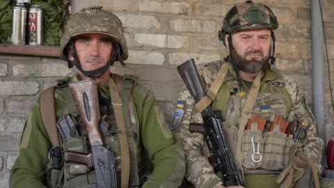  Two Ukrainian soldiers wearing helmets and carrying guns look into the camera lens