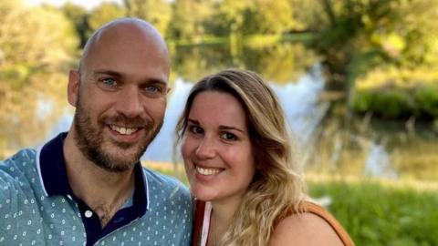 A couple smile at the camera. A man with a light beard wearing a light blue patterned polo shirt is to the left of the picture. A woman with shoulder length blonde hair stands beside him.