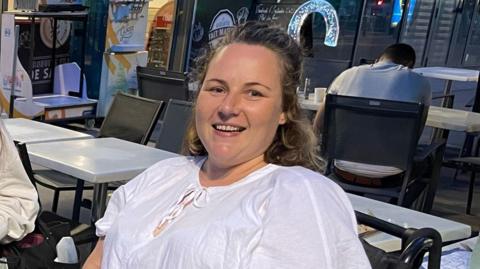 Sam Jennings, who has long brown hair tied back from her face and is wearing a white top. She is looking into the camera and smiling. There are tables and chairs behind her